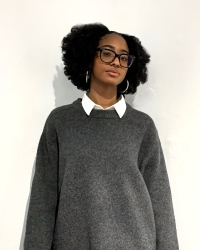 headshot of Black woman with black glasses standing in front of a white wall wearing a gray sweater and a white collar.