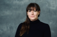 headshot of white woman with brown hair and bangs, wearing a black turtleneck and standing in front of a gray wall. 