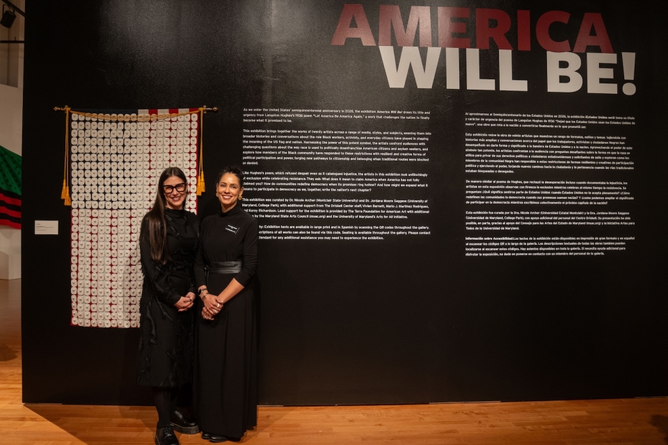 Two women stand in front of a piece of art and in front of an exhibition sign that says America Will Be! They are smiling.