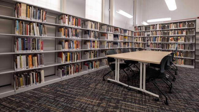 Wide shot of academic library with books on five levels of shelves at a right angle. Conference table in the middle of the image with 8 chairs.